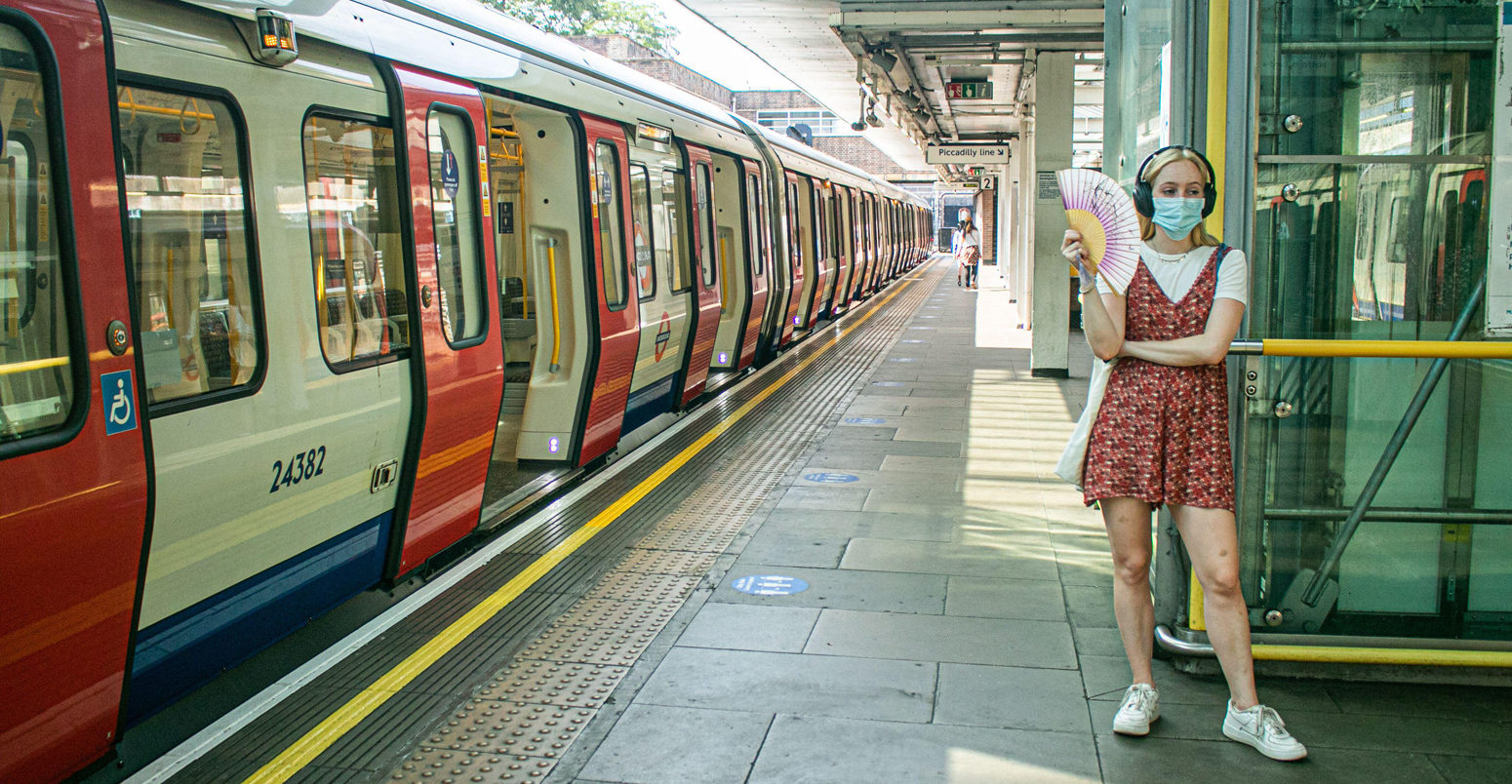 A passenger wearing a facemask standing on the London Underground platform using a fan to cool down from the heat and humid conditions during the August heatwave.
