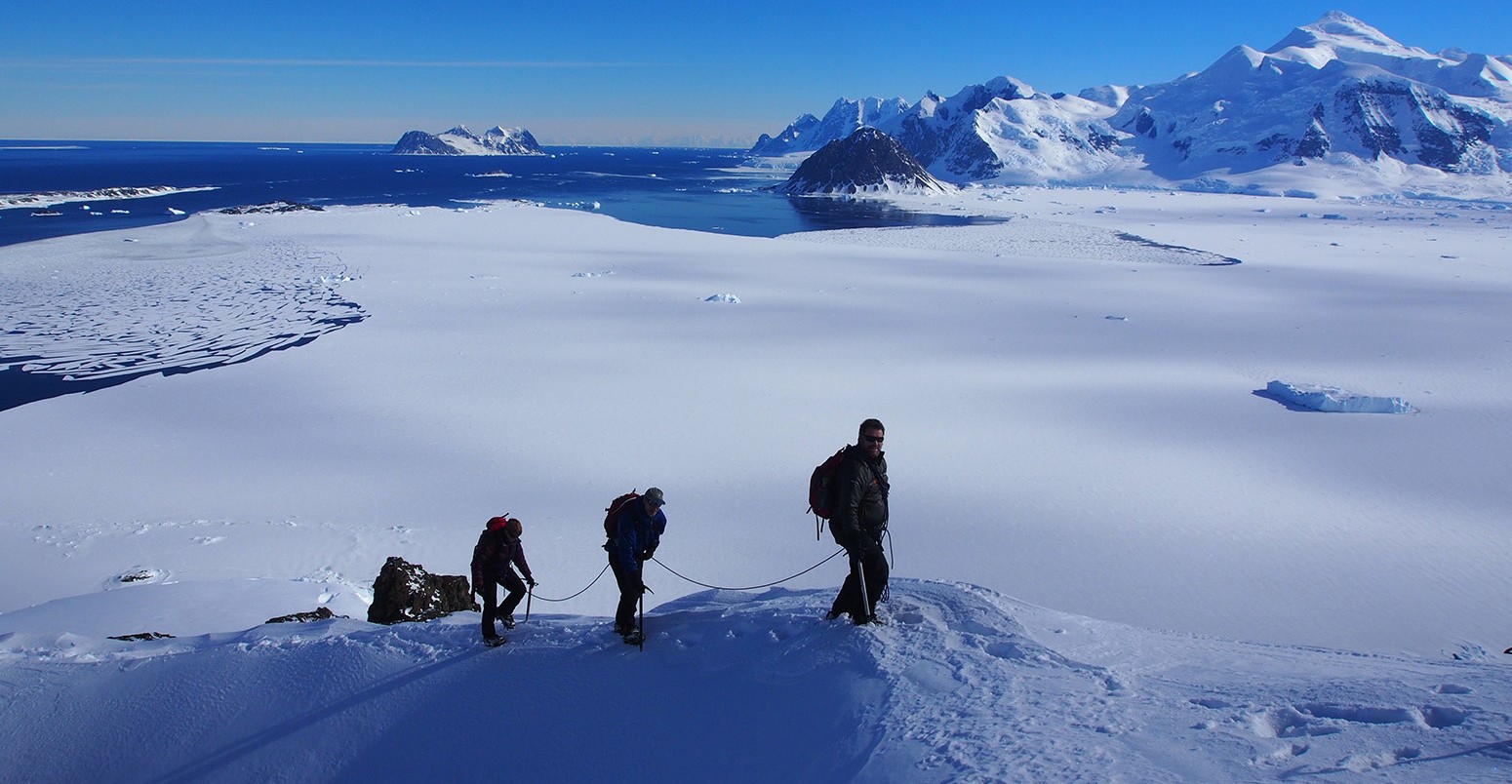 Researchers walking up a snowy ridge in a polar region overlooking the ocean