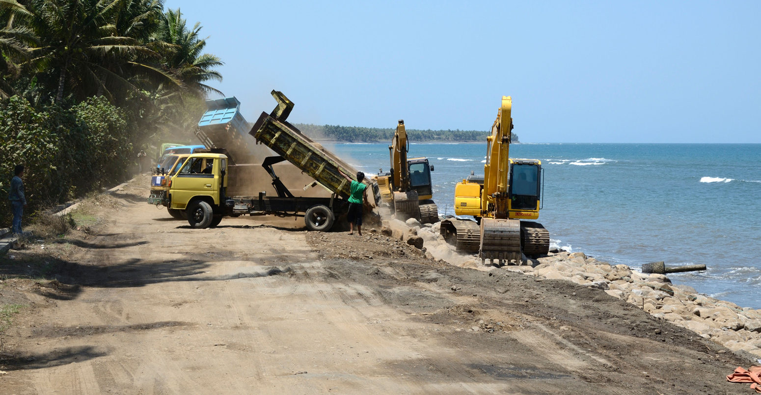 Trucks tipping boulders on to beach to build a new breakwater to prevent further coastal erosion. Lombok, Indonesia.