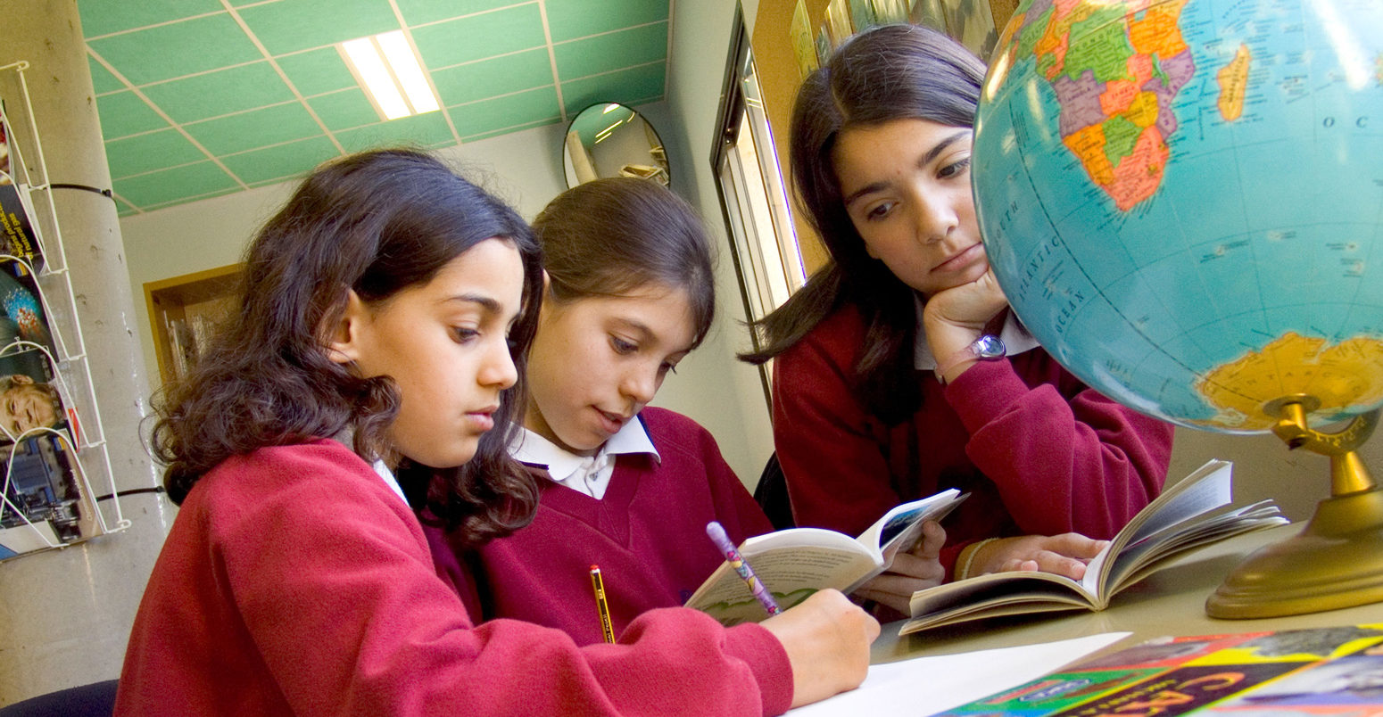 Three junior school girls studying the globe atlas for geography studies in school library_A0NP28